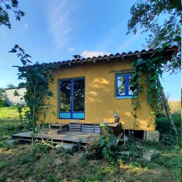 La imagen muestra una pequeña casa amarilla con tejado de tejas y ventanas de marco azul, rodeada de exuberante vegetación y un sereno cielo azul.