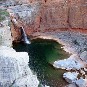 La imagen representa un entorno natural sereno, con una pequeña cascada o arroyo que desemboca en una tranquila masa de agua, rodeada de exuberante vegetación y un cielo azul despejado.