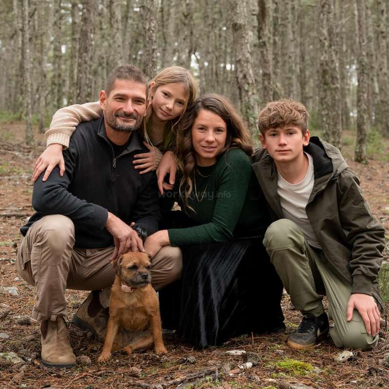 Une famille de quatre personnes, composée d'un homme, d'une femme, de deux enfants et d'un petit chien brun, pose joyeusement ensemble dans une forêt, entourée de grands arbres.