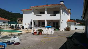 A imagem mostra uma casa com um grande quintal, com piscina e área de pátio, cercada por árvores e arbustos, com uma entrada de automóveis que leva à casa. A casa tem um design moderno.