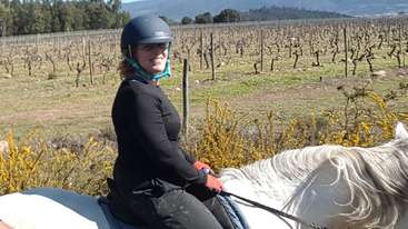 A imagem mostra uma pessoa montada em um cavalo, com um corpo de água ao fundo. A pessoa está usando um capacete e montando um cavalo, com uma cerca visível à distância.
