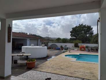 A imagem mostra uma cena serena no quintal, com piscina, banheira de hidromassagem, móveis de pátio e uma garagem, em um céu nublado com árvores ao fundo.