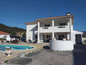 A imagem mostra uma casa branca de dois andares com telhado vermelho, com piscina e área de pátio, tendo como pano de fundo árvores e um céu azul claro.