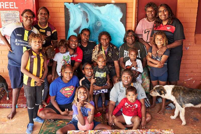 Un grand groupe d'adultes et d'enfants aborigènes australiens posent ensemble, souriants, devant un mur de briques et des œuvres d'art colorées. Un chien se tient à droite.