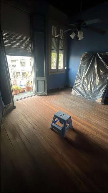A mostly empty room with dark blue walls, wooden floors, a blue step stool, ceiling fan, covered object, open door to a balcony with plants.