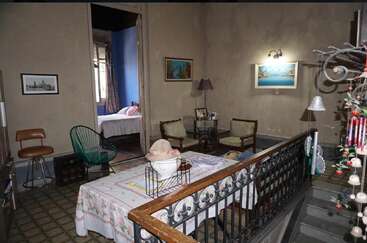 This image shows a cozy, vintage living area with mismatched furniture, wall art, and a table with a cover. An adjacent bedroom is visible through the doorway.