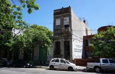 A two-story old building stands between trees and parked cars. Brightly lit by the sun, it features a faded facade and a large white advertisement board.