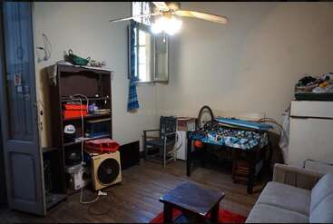 This cozy room features wooden floors, a ceiling fan, bookshelf, foosball table, open window with blue curtain, couch, small table, and eclectic decor.