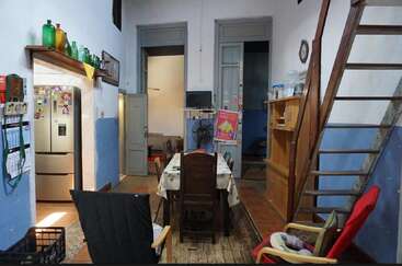 This cozy dining room features a wooden table, mismatched chairs, a wooden staircase, kitchen with fridge, shelves, colorful bottles, and blue-and-white painted walls.
