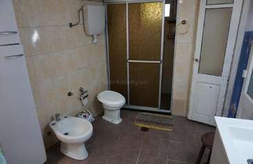 This is a spacious, tiled bathroom featuring a bidet, toilet, shower with frosted sliding doors, cabinet, white sink, small stool, and a patterned floor mat.