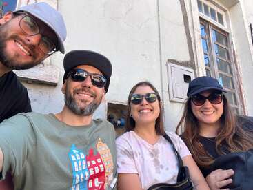 Four friends, all wearing sunglasses and smiling, pose for a selfie outside a weathered building with a window. They seem happy, relaxed, and enjoying the day.