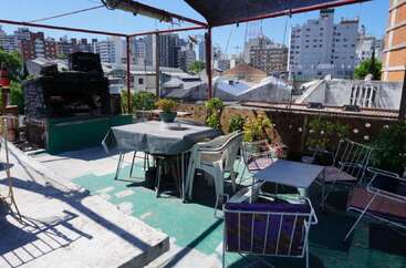 Une terrasse sur le toit avec des tables, des chaises et des plantes, à l'ombre d'un auvent. Il y a un grill en briques dans le coin et des bâtiments de la ville à l'arrière-plan.