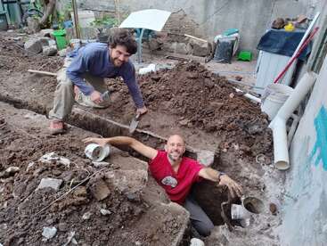 Two men are digging a hole in the ground, surrounded by dirt and debris, with one man sitting in the hole and the other kneeling beside it.