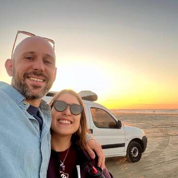 A smiling couple takes a selfie on a sandy beach at sunset, standing in front of a white car with the ocean and clear sky in the background.