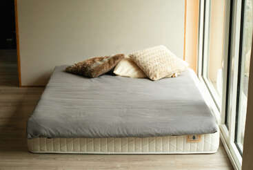 Une chambre à coucher minimaliste comprend un matelas bas posé sur le sol avec un drap gris, trois oreillers texturés et de grandes fenêtres laissant entrer la lumière naturelle. Atmosphère douillette.