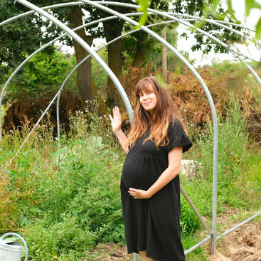 Eine schwangere Frau in einem schwarzen Kleid steht in einem grünen Garten, hält ihren Bauch und winkt, umgeben von Metallbögen, Bäumen und überwucherten Pflanzen.