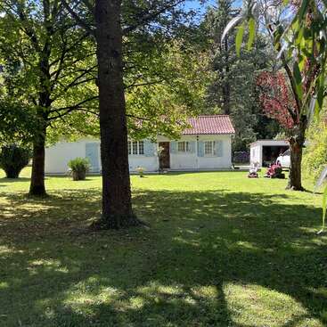 A charming white house with blue shutters sits in a lush, green yard shaded by tall trees. A garage and lawn equipment are visible nearby.