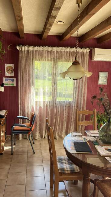 A cozy dining room with wooden beams, red-striped walls, sheer curtains, round glass table, padded chairs, sunlight streaming in, wall art, desk, and plants.