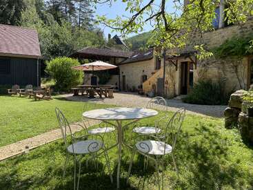 L'image représente une arrière-cour sereine, avec une table et des chaises blanches, un parasol marron et une maison en pierre, entourée d'une végétation luxuriante et d'un ciel bleu clair.