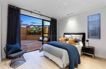 Um quarto moderno apresenta uma cama aconchegante, decoração elegante, cortinas escuras e uma porta de vidro deslizante que se abre para um deck de madeira com palmeiras e assentos confortáveis.