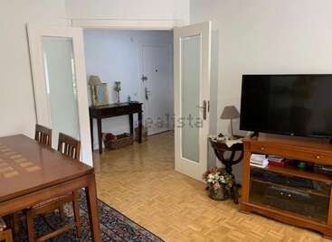 This cozy living room features a wooden dining table, TV stand, television, side table with lamp, decorative flowers, rug, and an open doorway to a hallway.