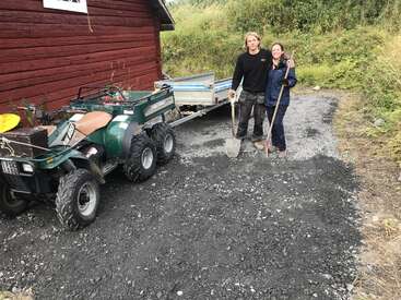 Deux personnes se tiennent debout, souriantes, une pelle à la main, sur une allée en gravier près d'un bâtiment en bois rouge. Un quad avec une remorque est garé à côté d'elles, au milieu de la verdure.