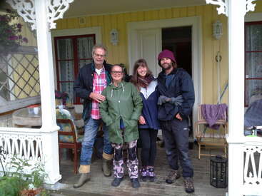 The image depicts three individuals standing on a porch, dressed in casual attire, with a house and outdoor furniture visible in the background.