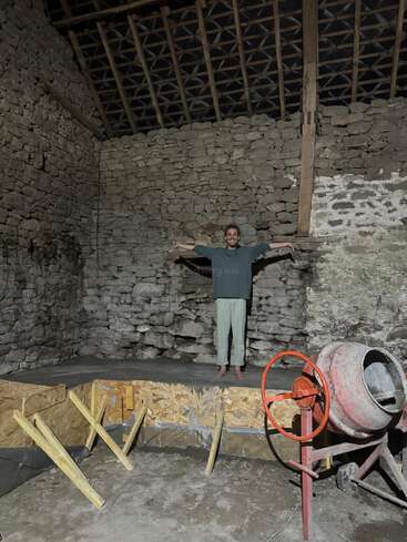 Una persona está de pie, descalza, sobre una plataforma de hormigón recién construida en el interior de un edificio rústico con paredes de piedra. En primer plano se ven herramientas de construcción, entre ellas una hormigonera.