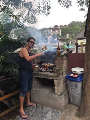 A smiling man in sunglasses barbecues meat outdoors, holding tongs and a drink. Smoke rises from the grill; a beer bottle stands nearby on a brick barbecue.