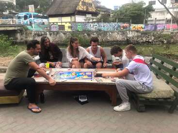 Six people sit outdoors around a table playing a board game, enjoying each other's company. Graffiti decorates the wall behind them, creating a lively urban atmosphere.