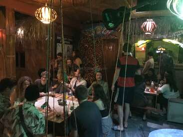 A group of people gathers inside a cozy, dimly lit bar or café with wooden decor, colorful lamps, and posters, enjoying conversation, drinks, and a relaxed atmosphere.