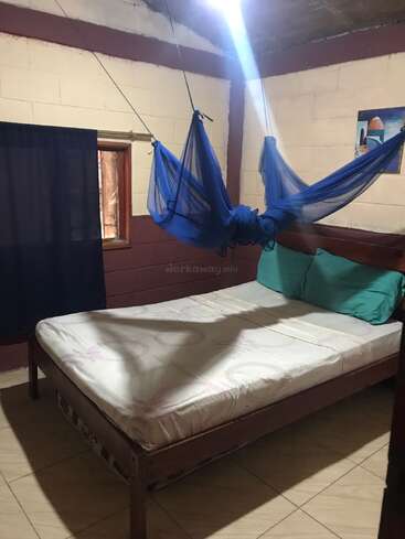 A simple room with a wooden bed, white mattress, teal pillows, blue mosquito net hanging above, small window, tiled floor, painting, and partial sunlight.