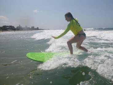 Una mujer con una camiseta amarilla y un bikini verde surfea sobre una tabla verde en pequeñas olas cerca de la orilla bajo un cielo soleado.