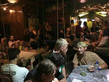 Una animada reunión social en interiores con jóvenes adultos conversando, bebiendo y riendo. Iluminación cálida, ambiente informal, guirnaldas de luces, bar al fondo y decoraciones eclécticas.