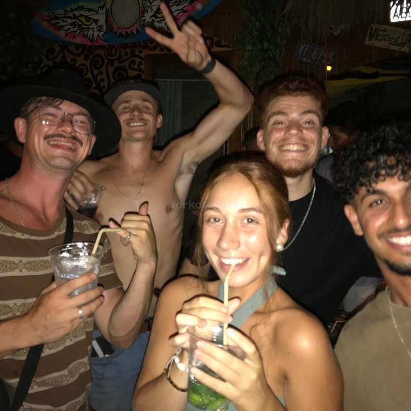 Un grupo de cinco jóvenes adultos sonríen y beben en una animada fiesta, uno de ellos está sin camiseta, todos parecen felices, relajados y llenos de energía.
