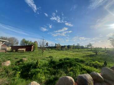 Un hermoso día soleado en un prado salpicado de árboles, con un cobertizo de madera, rocas en primer plano y un cielo azul brillante con nubes.