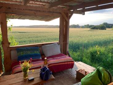 Eine gemütliche Holzveranda mit einer bunten Bank bietet einen Blick auf eine ruhige, sonnenbeschienene Wiese. Blumen, Kissen, Taschen und ein Getränk sorgen für eine friedliche, einladende Oase.