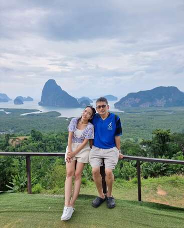 La imagen muestra a un hombre y una mujer posando para una foto en un balcón con vistas a un paisaje exuberante con montañas lejanas y agua, en un cielo nublado.