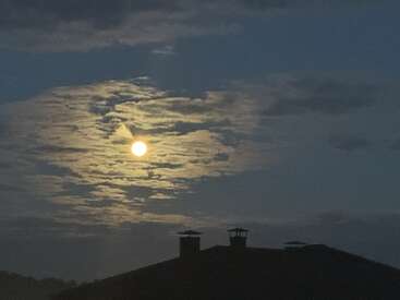 A bright full moon shines through wispy clouds in a dark evening sky, with the silhouette of a rooftop and twin chimneys visible below. Peaceful night.