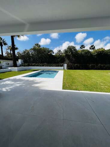 Das Bild zeigt einen modernen Hinterhof mit einem kleinen, rechteckigen Pool, umgeben von grünem Gras, Palmen, blauem Himmel, weißen Wolken und einer schlichten Terrasse.