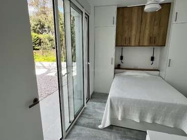 Dieses Bild zeigt ein kleines, helles Schlafzimmer mit einem Doppelbett, eingebautem Stauraum, großen Glasschiebetüren und moderner Einrichtung. Der Raum öffnet sich zu einem Garten.