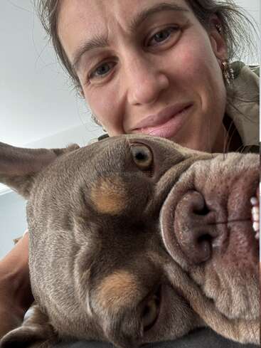 Eine Frau lächelt in die Kamera, während sie ihren entspannten, braunen Hund streichelt. Das Gesicht des Hundes ist ganz nah an der Linse und wirkt ruhig und zufrieden.