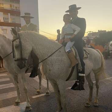 Das Bild zeigt einen Mann und ein Kind, die auf einem weißen Pferd auf einer Straße reiten. Hinter ihnen stehen zwei Männer in traditioneller Kleidung vor einer Stadtkulisse.