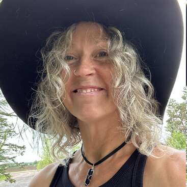 A woman with curly blonde hair smiles outdoors. She wears a large black hat, black tank top, nose ring, and black pendant necklace. Trees are in the background.