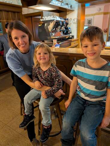 Una mujer y dos niños sonrientes se sientan en los taburetes de una acogedora cafetería, con una cálida decoración de madera, una cafetera y vasos a la vista detrás de ellos.