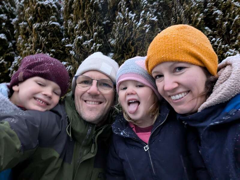 Una feliz familia de cuatro miembros posa al aire libre con ropa de invierno. Dos adultos y dos niños sonríen, con un niño sacando juguetonamente la lengua, rodeados de árboles nevados.