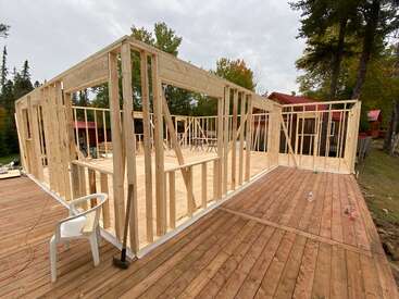 Une ossature de bâtiment en bois, en cours de construction, se dresse sur une terrasse en bois à l'extérieur, entourée d'arbres. Des outils de chantier, des bouteilles et une chaise en plastique blanche se trouvent à proximité.