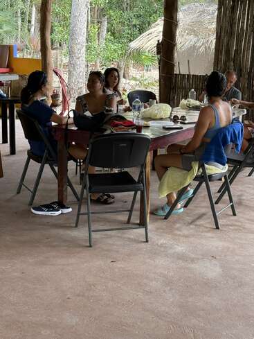 Five people sit around a table outdoors, enjoying food and conversation. The area is rustic, with trees, wooden beams, and natural surroundings visible in the background.