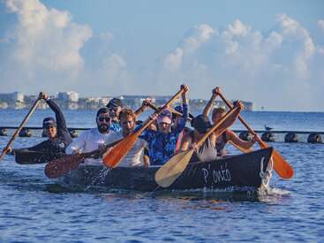 A team of people energetically paddle a canoe on calm blue water, working in unison. In the background, buildings and fluffy clouds are visible.