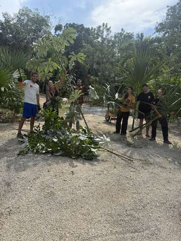 Sechs lächelnde Menschen im Freien halten große, abgeschnittene Äste und Werkzeuge und stehen auf Kies, umgeben von Grünzeug. Sie scheinen gemeinsam an einem Garten- oder Landschaftsbauprojekt zu arbeiten.